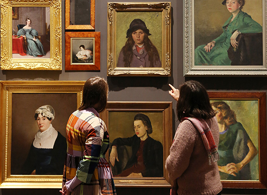 Two women in colourful clothing are looking at a wall covered in portrait paintings