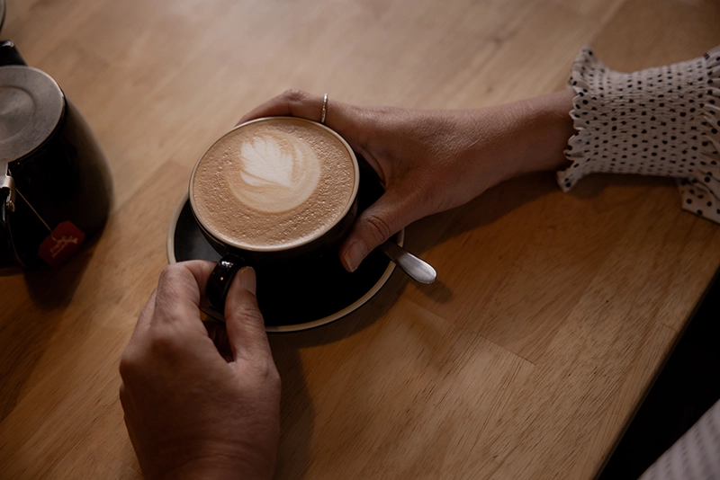 Hands holding a cup of coffee with latte art on a wooden table.