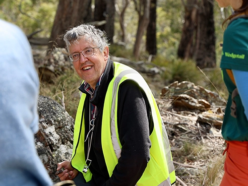 Person in high-visibility vest speaking with others in a bushland setting.