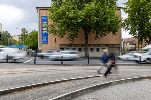 Street view of the Tasmanian Museum and Art Gallery with blurred traffic and pedestrians passing by.