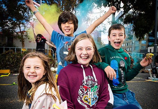 Four young Lift Off! Festival attendees smile at the camera while colourful smoke billows behind them